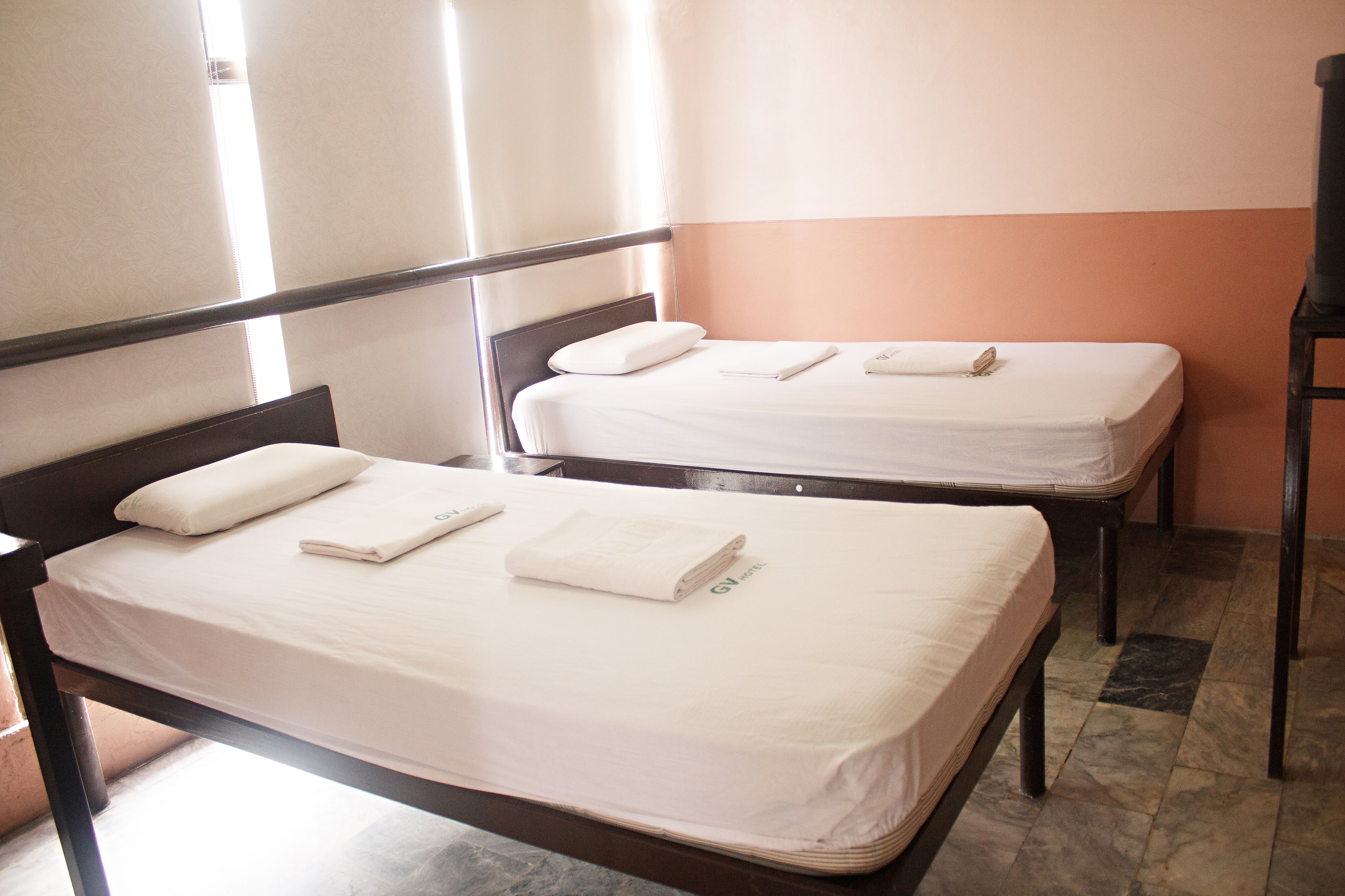Dos camas individuales con sábanas y almohadas blancas, un cabecero de madera y piso de baldosas.