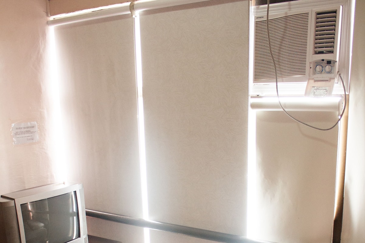 A hotel room with a bed, a TV on a stand, a window with blinds, and an air conditioning unit.