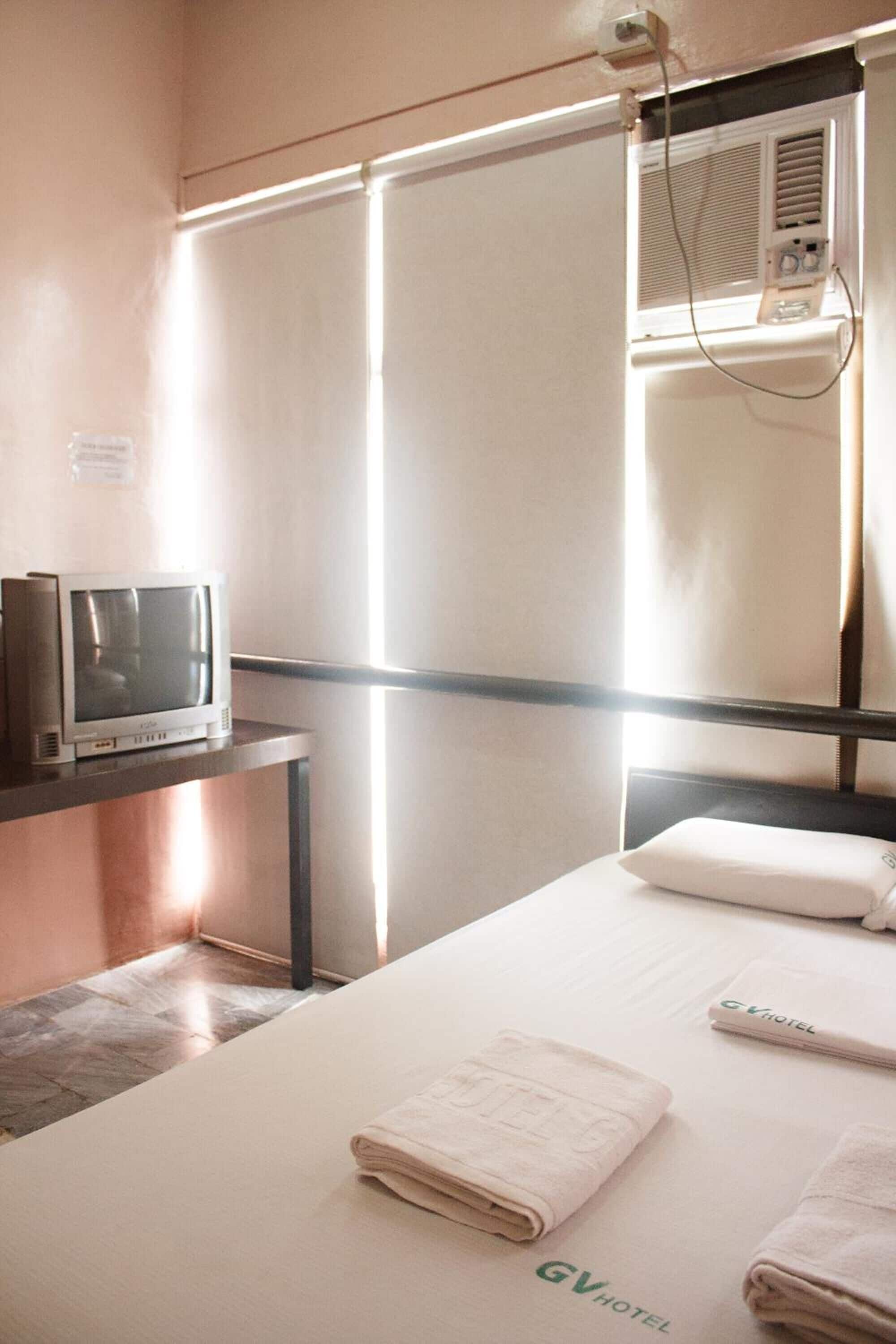 Una habitación de hotel con una cama, un televisor sobre un mueble y una ventana con persianas.