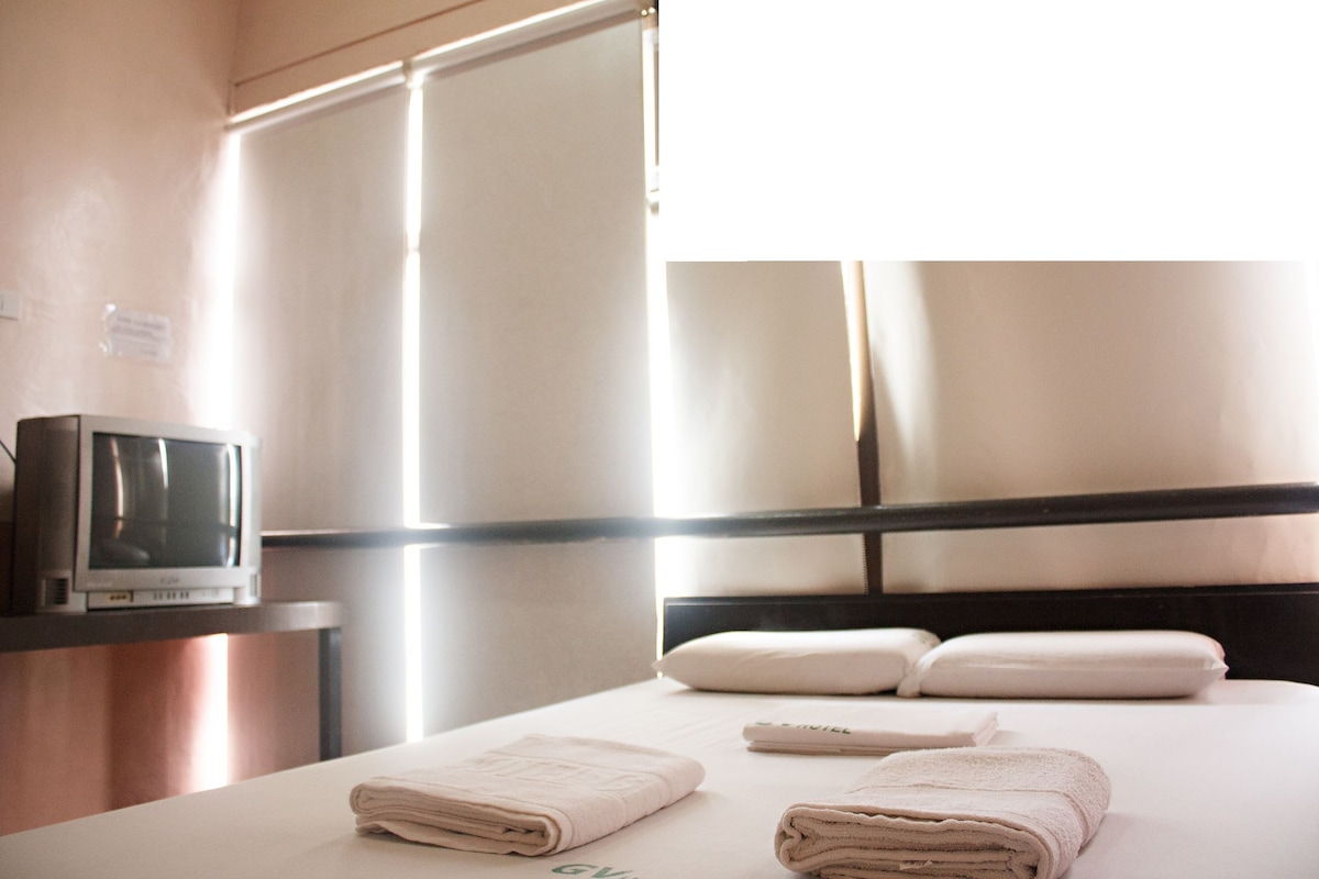 A hotel room with a bed, pillows, a TV on a stand, and a window.