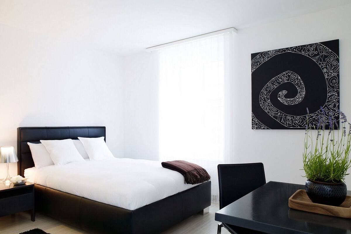 A modern bedroom with a black bed, white bedding, a small bedside table with a lamp, a dark wooden side table, a black chair, a black dining table, a potted plant, and a large abstract artwork on the wall.