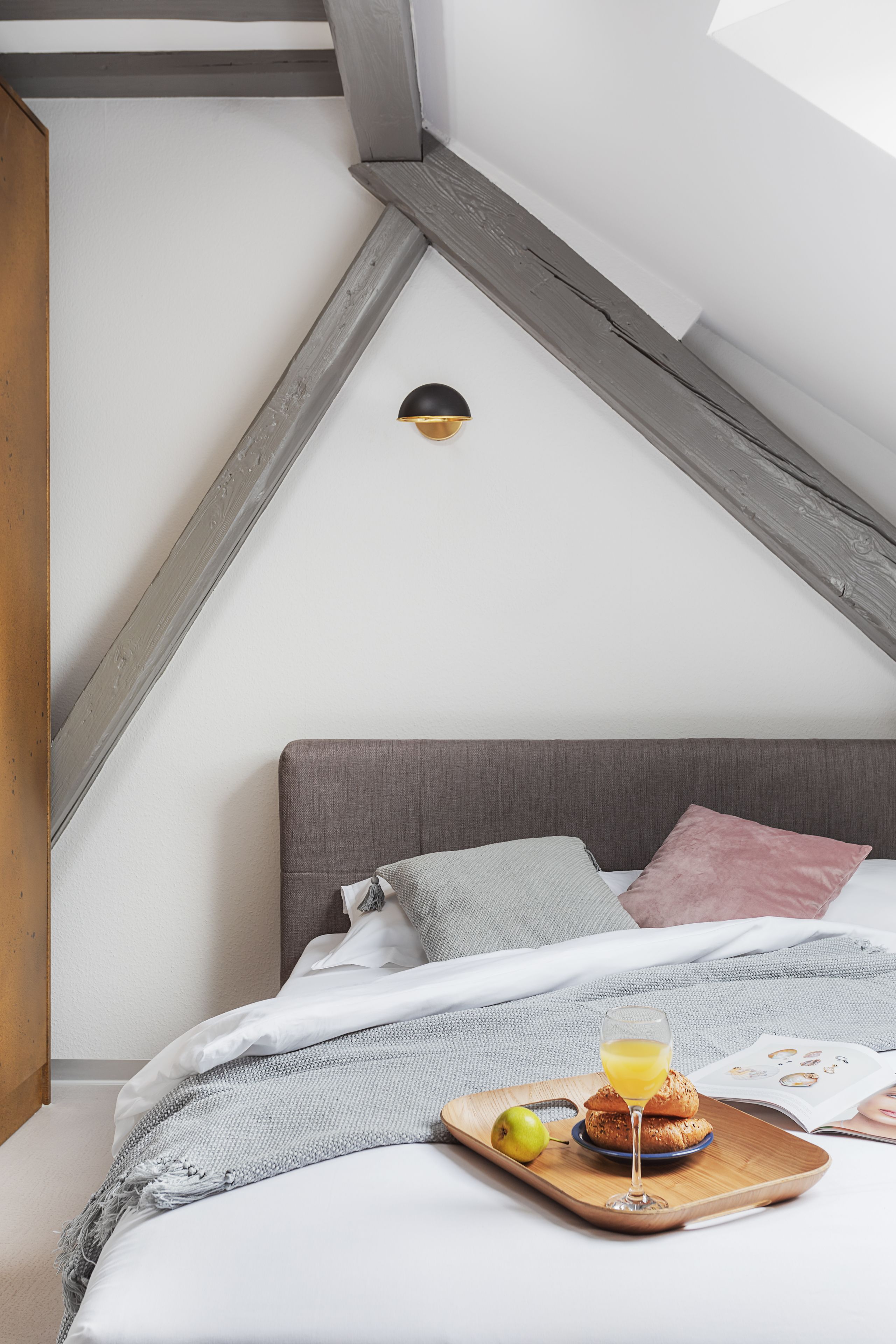 Ein Schlafzimmer mit schrägem Dach, einem Holzkopfende und einem Frühstücksbrett mit Muffins, einem Glas Orangensaft und einem Buch auf dem Bett.