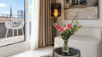 Un salon moderne avec un canapé blanc, une table ronde noire ornée d’un vase de fleurs, un lampadaire et une vue sur les immeubles à travers une grande fenêtre.