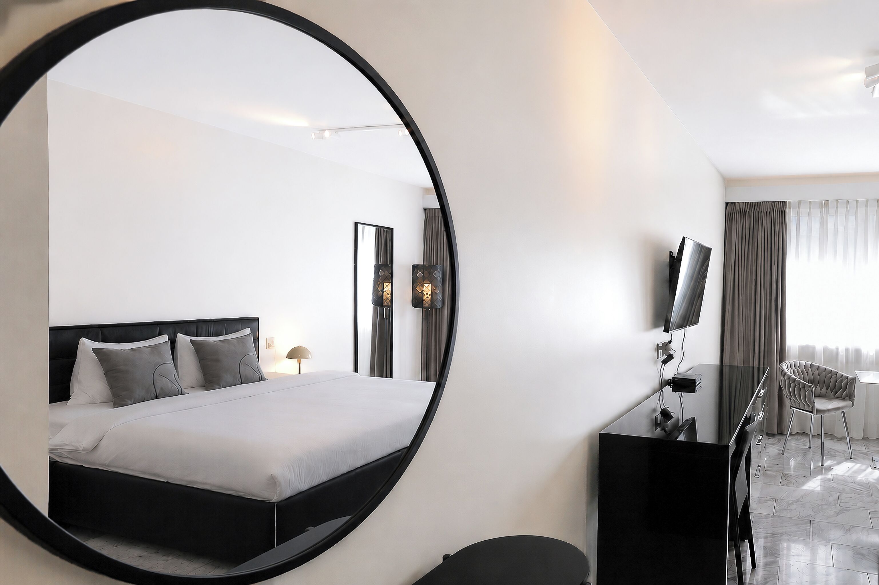 Habitació d'hotel moderna amb un gran mirall rodó, un llit amb roba de llit gris, una taula de nit amb un televisor de pantalla plana i una cadira.
