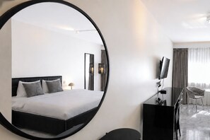Een moderne hotelkamer met een grote ronde spiegel, een bed met grijze beddengoed, een bureau met een flatscreen televisie en een stoel.