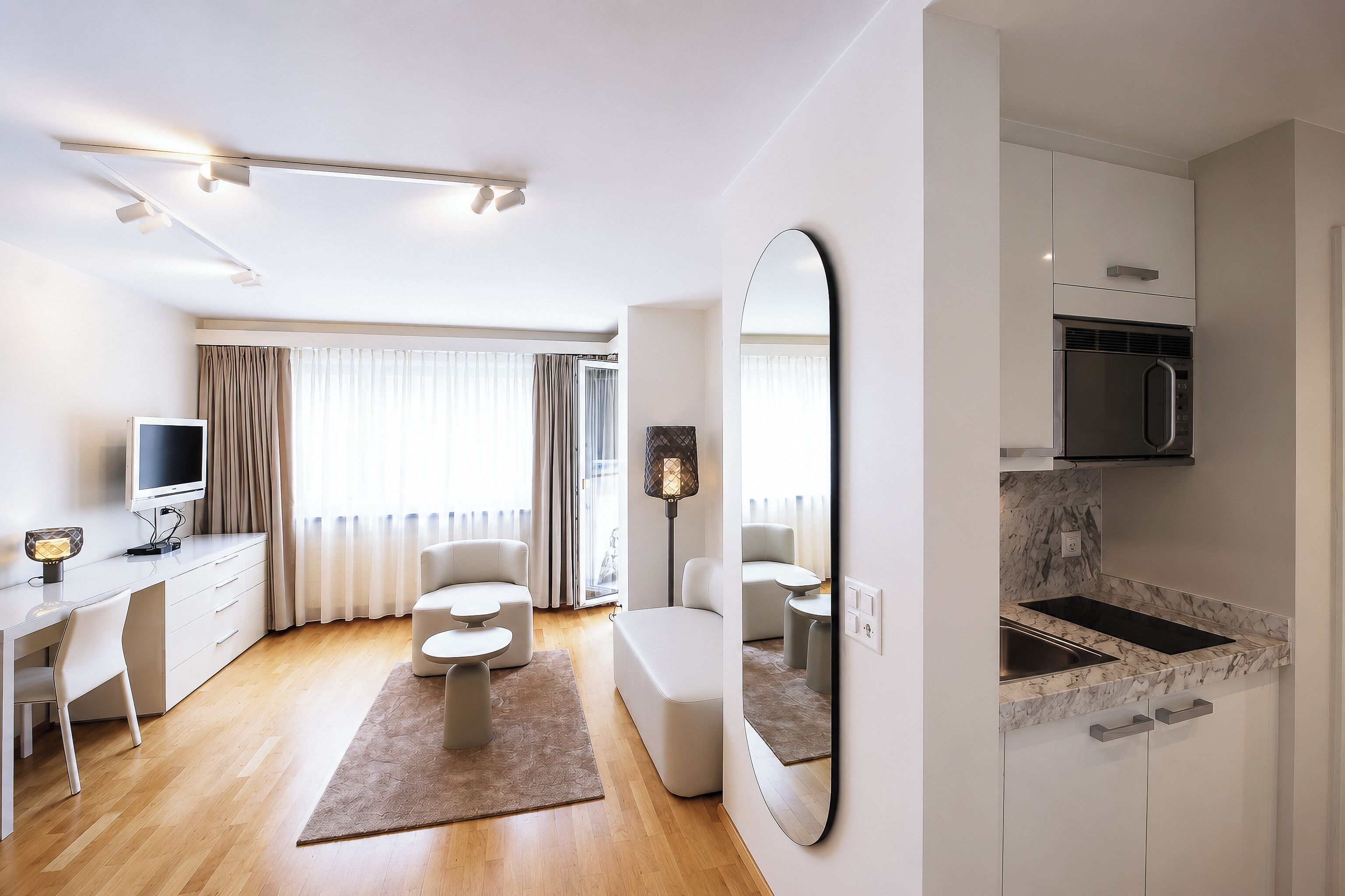 A modern hotel room with a flat-screen TV, a sofa, a dining area, a kitchen with a built-in oven, and a large mirror.
