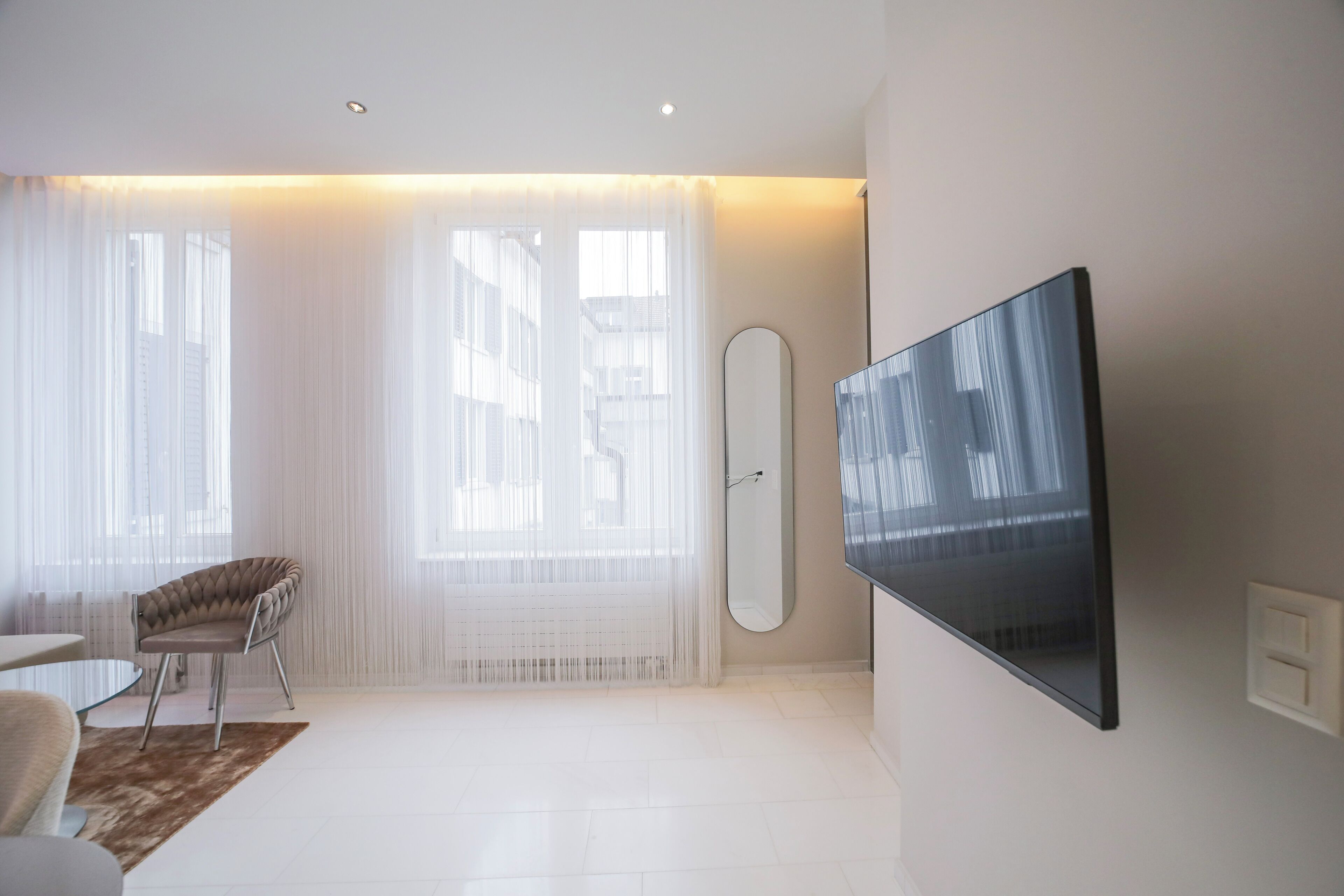 Un salon moderne avec un téléviseur à écran plat fixé au mur, un fauteuil confortable et une grande fenêtre ornée de rideaux légers.