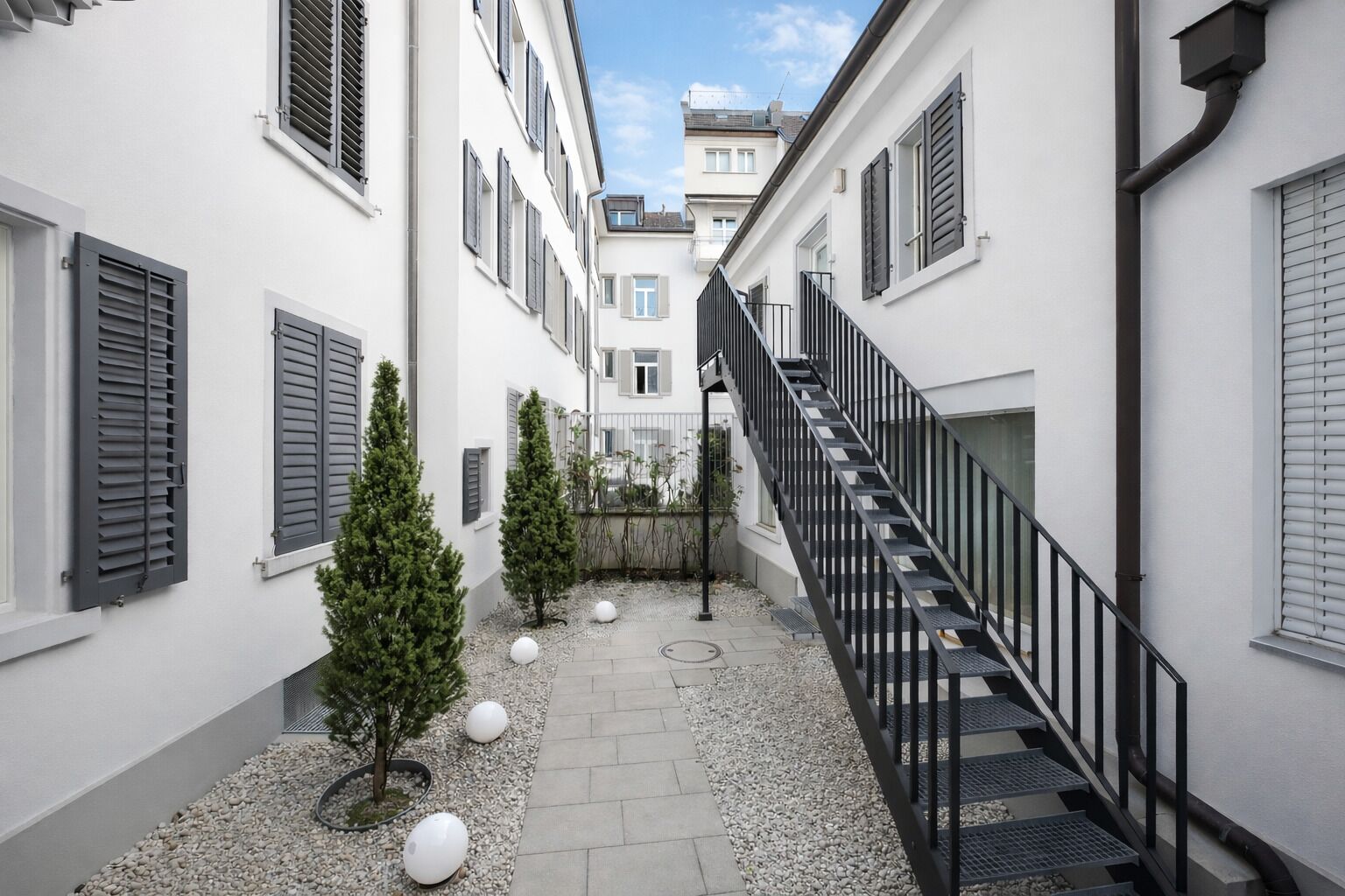 Ein schmaler Innenhof mit einer Treppe, die zu einer oberen Ebene führt, umgeben von Gebäuden mit Fensterläden und kleinen Bäumen.