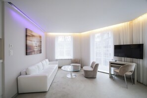 Una sala de estar moderna con un sofá, una mesa redonda de centro, sillas, un televisor de pantalla plana y ventanas amplias con cortinas.