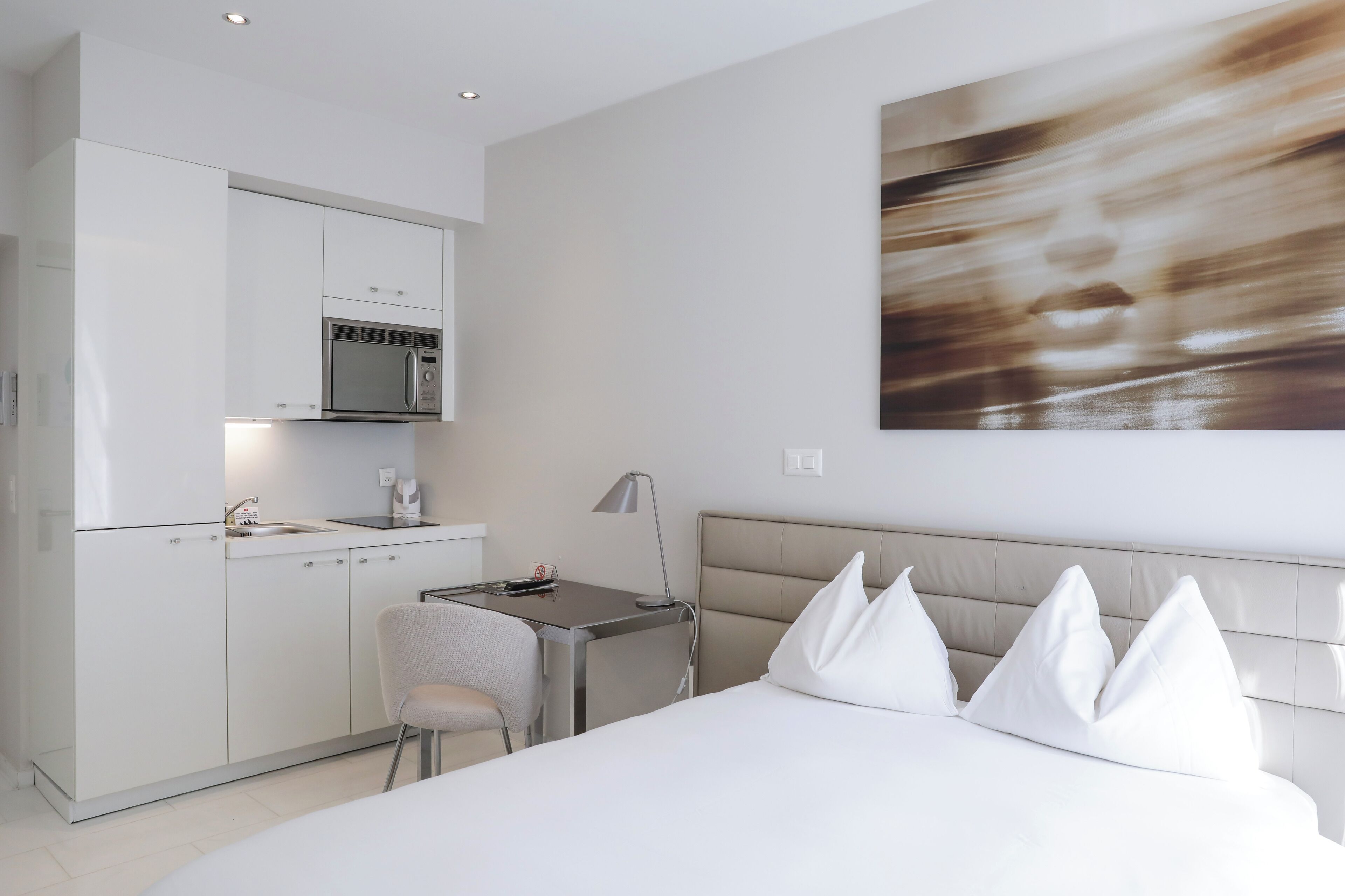 A modern hotel room with a kitchenette, a bed with white pillows, and a large abstract painting on the wall.