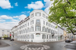A multi-story building with a corner entrance, a visible logo on the facade, and a parking area with marked spaces. - VISIONAPARTMENTS Zurich Gerechtigkeitsgasse (Zürich)