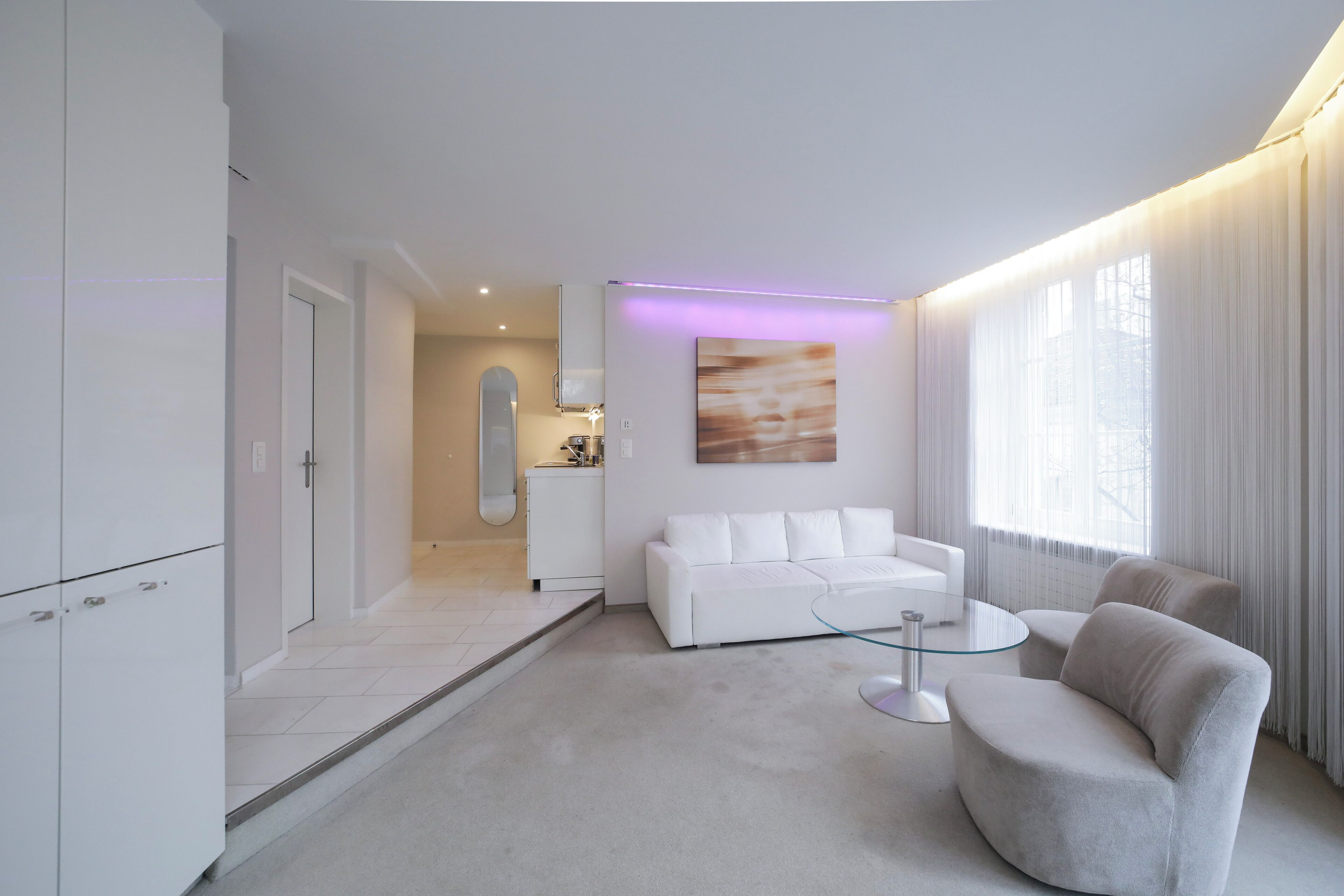 Habitació d'hotel moderna amb un sofà blanc, una taula de centre de vidre i una zona de menjador amb un armari blanc i un taulell de cuina.