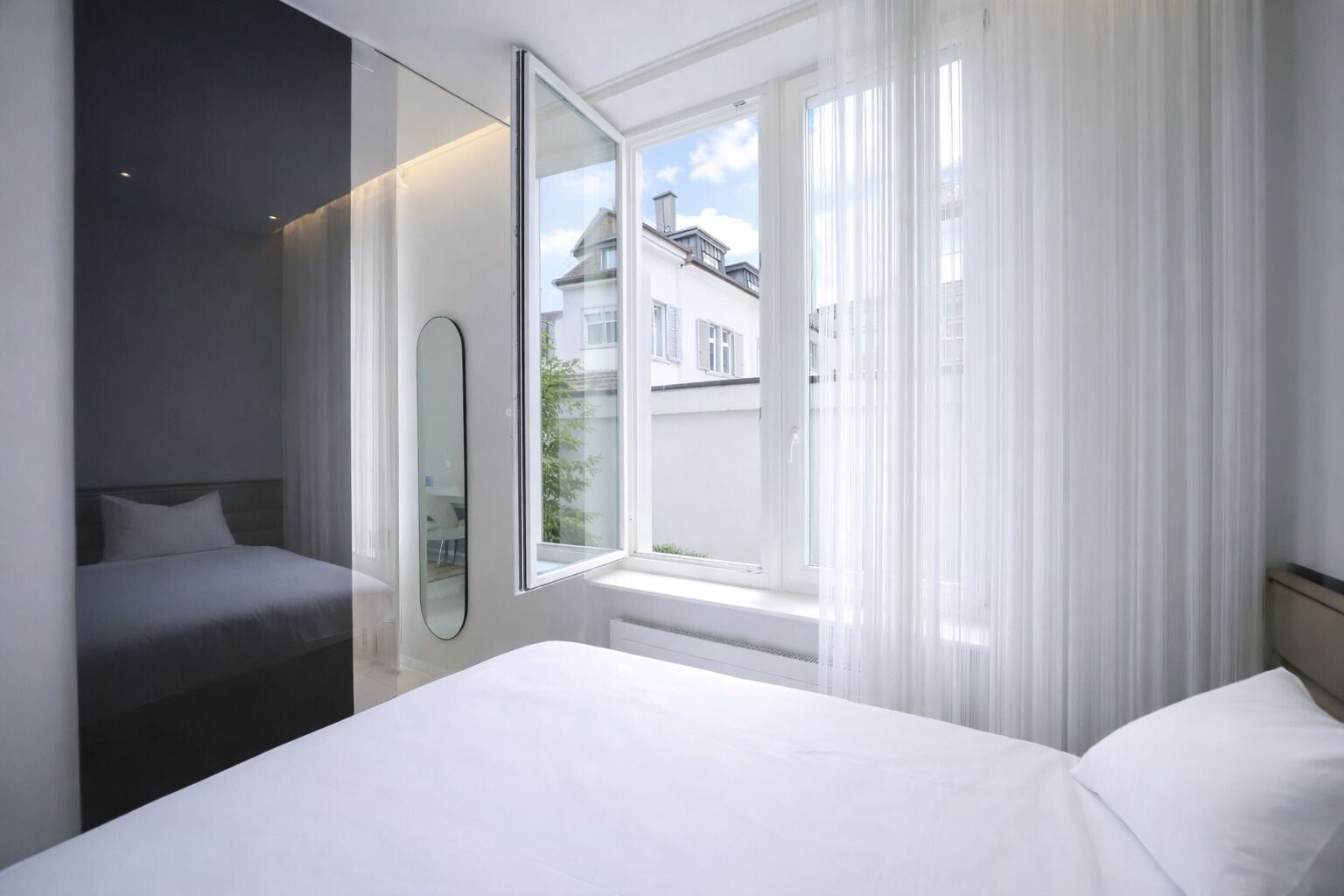 A modern hotel room with a large bed, a bedside table, a mirror, and a window with sheer curtains.