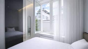 Une chambre d’hôtel moderne avec un grand lit, une table de chevet, un miroir et une fenêtre avec des rideaux légers.
