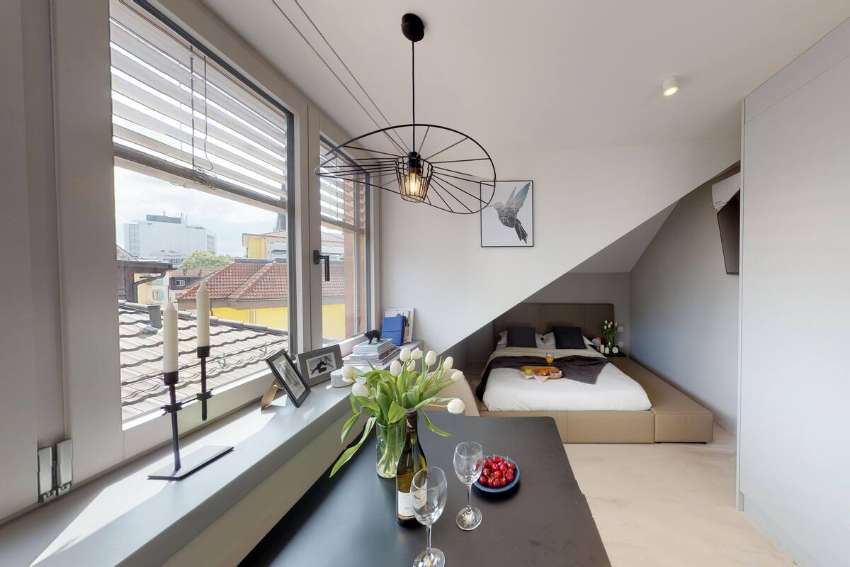 A modern bedroom with a bed, a desk with a vase of flowers, and a view of the city through large windows.