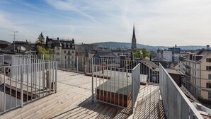 A rooftop terrace with a wooden deck and metal railing, overlooking a cityscape with buildings and a church spire. - VISIONAPARTMENTS Zurich Zweierstrasse (Zürich)