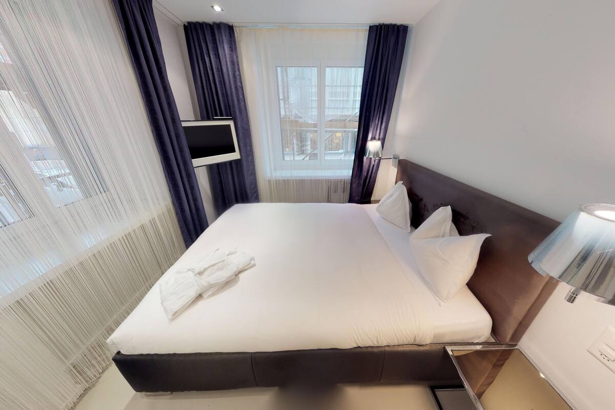 A hotel room with a large bed, a flat-screen TV mounted on the wall, and a window with curtains.