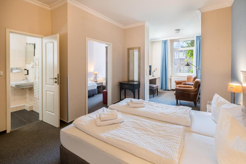 Une chambre d’hôtel avec deux lits, un coin salon comprenant une chaise et une table, une salle de bain avec un lavabo et un miroir, et une fenêtre avec des rideaux.