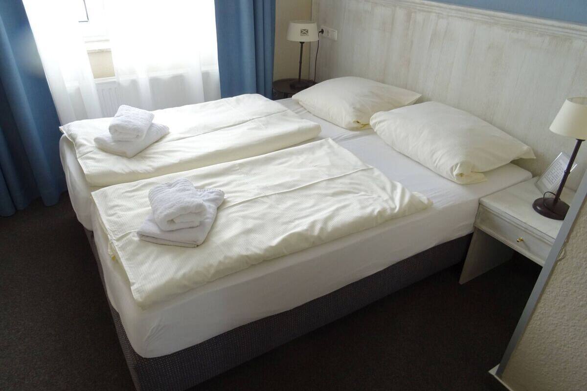 A double bed with white linens and towels, a bedside table with a lamp, and a window with blue curtains.