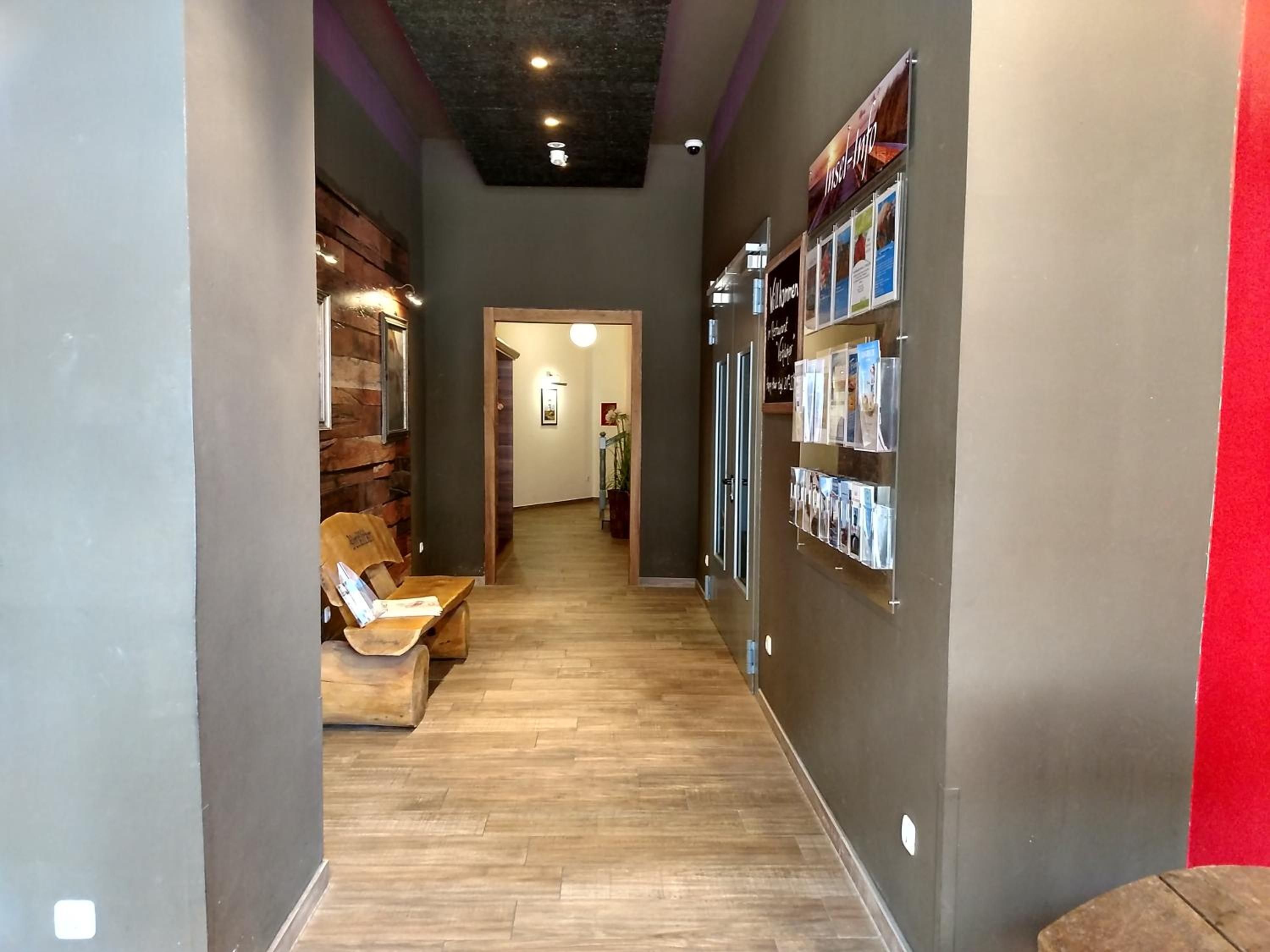 A hallway with a wooden floor, a seating area with chairs, and a wall with framed pictures and a bulletin board.