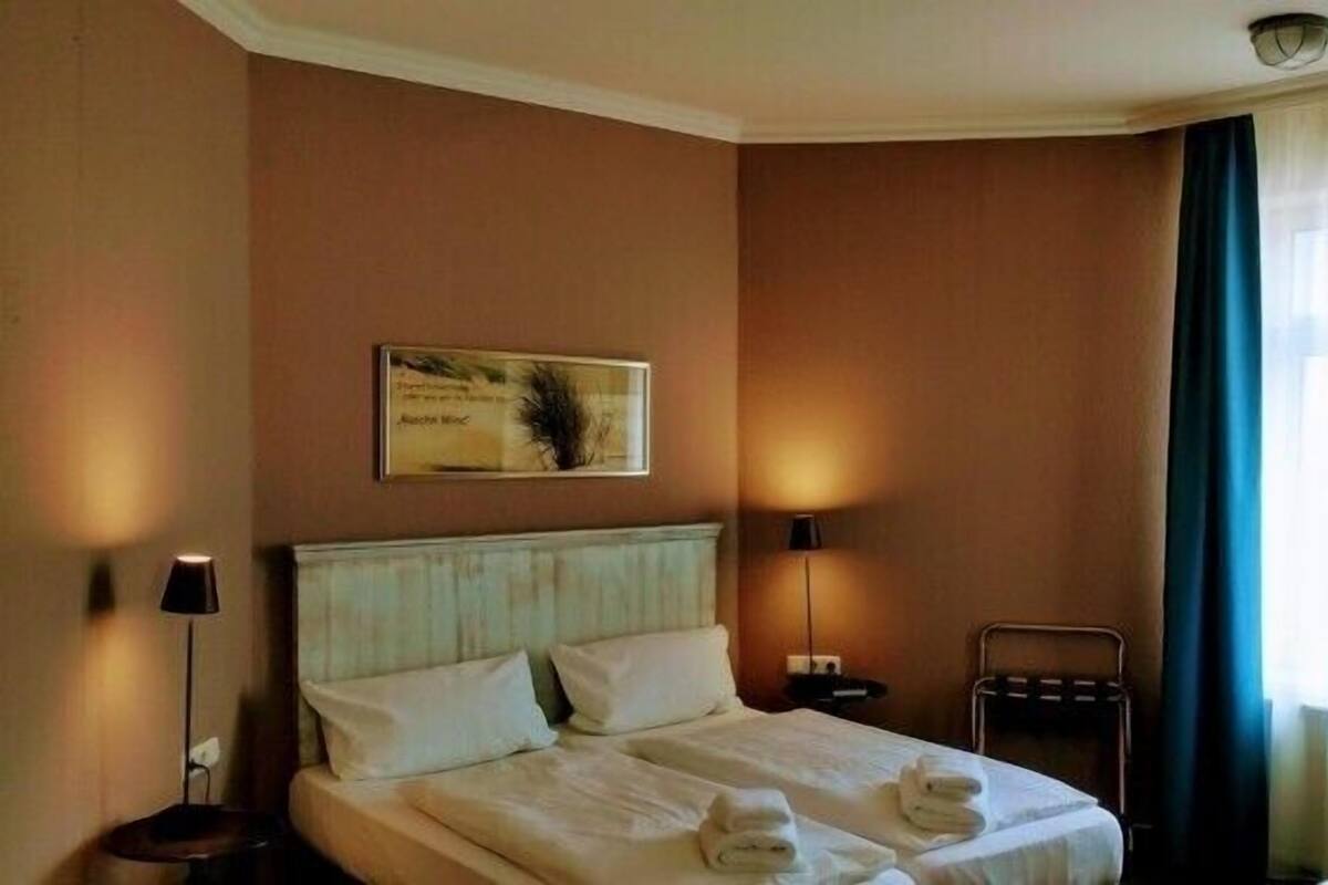 A hotel room with a bed, a bedside table, a lamp, a towel rack, and a window with curtains.