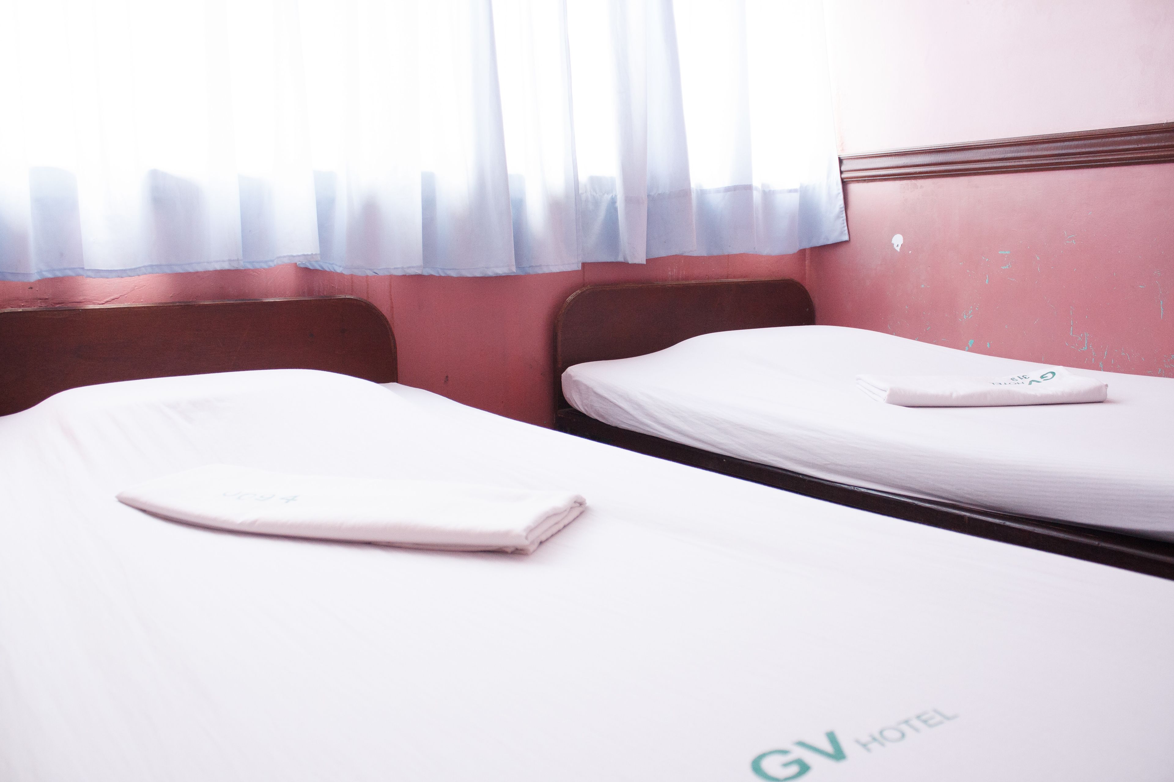 A hotel room with two single beds, a wooden headboard, and a window with sheer curtains.