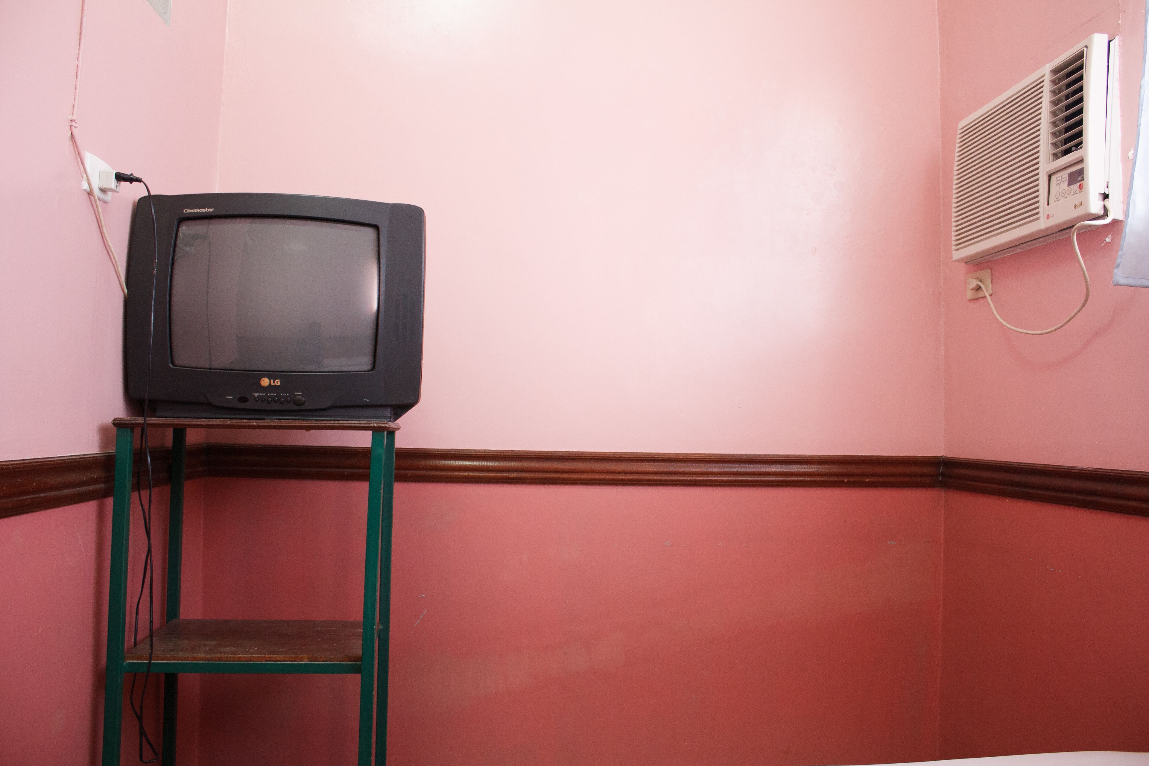 Un televisor sobre un estante de madera frente a una pared rosa con un equipo de aire acondicionado blanco.