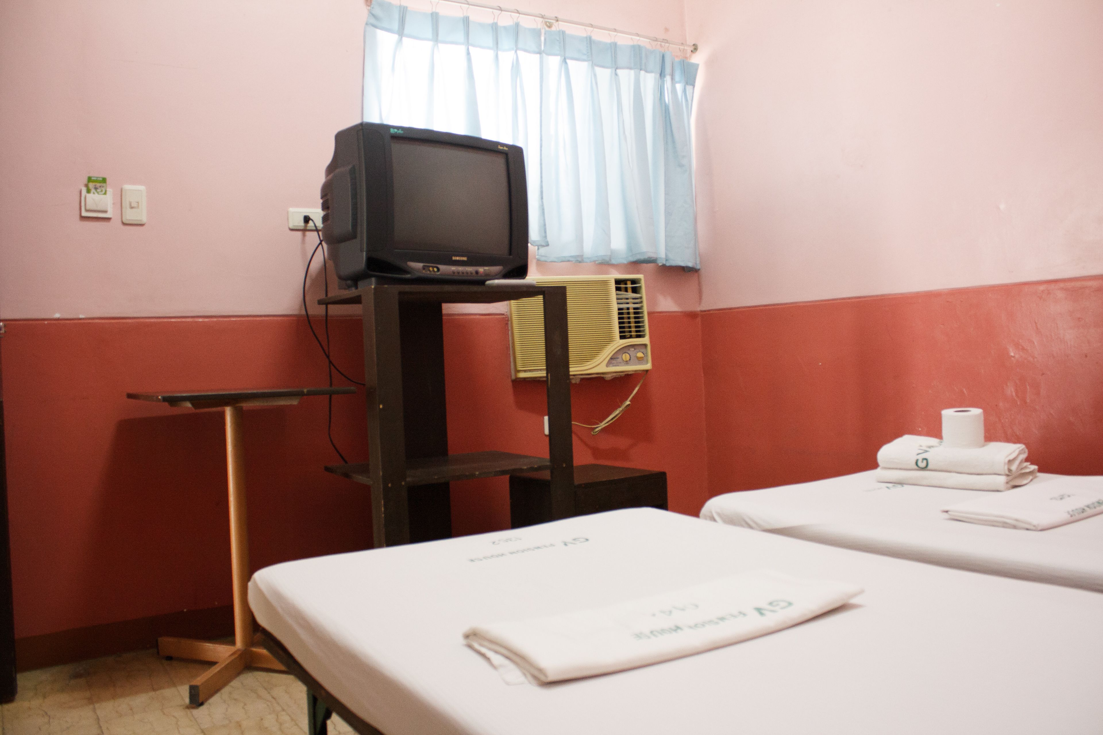A room with a television on a stand, an air conditioner, two beds with towels, and a window with curtains.