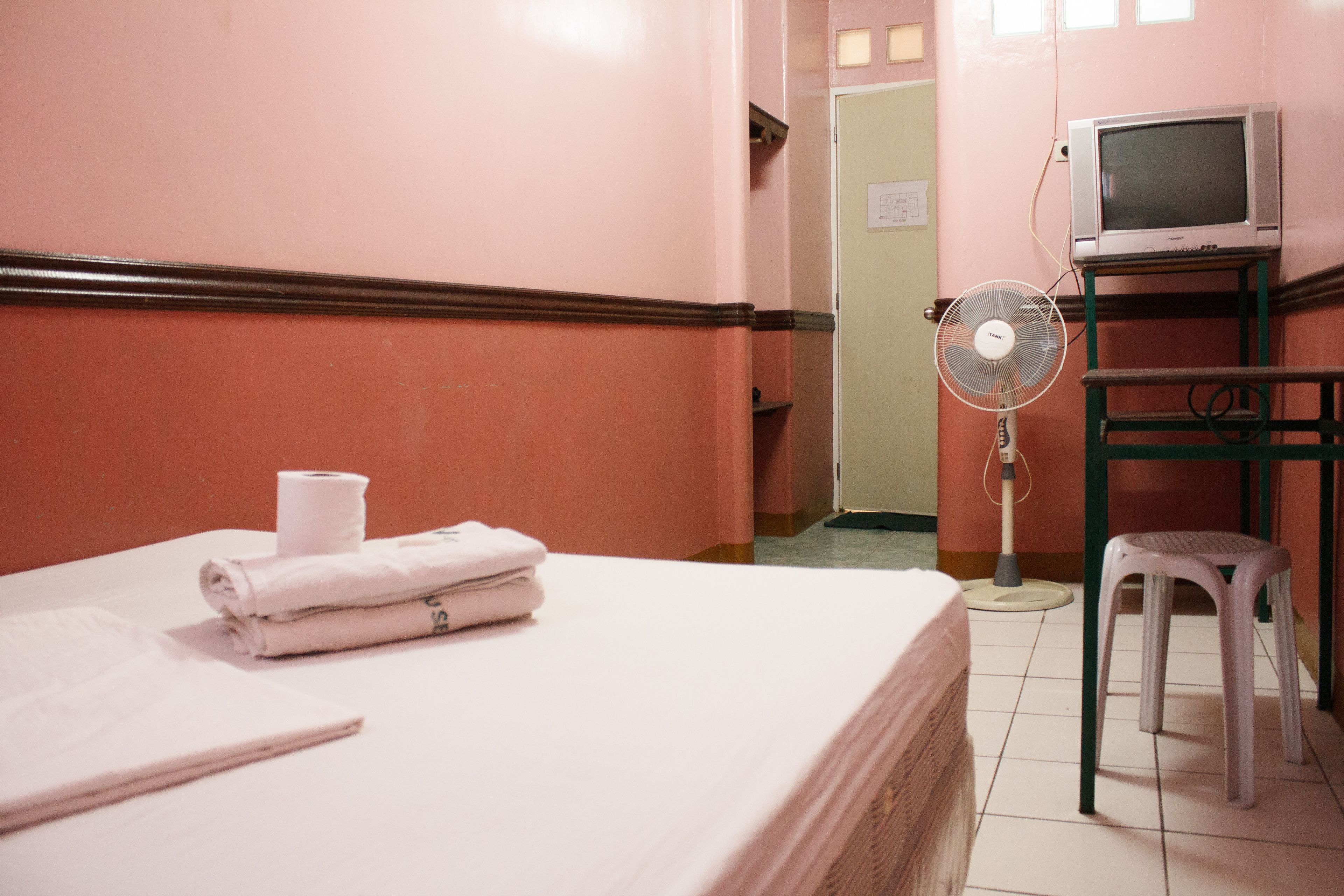 Una habitación de hotel con una cama, un ventilador, un televisor sobre un soporte y un taburete pequeño.