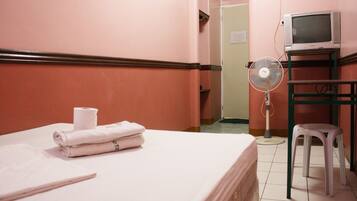 Una habitación de hotel con una cama, un ventilador, un televisor sobre un soporte y un taburete pequeño.