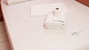 Een enkel bed met een witte matras, kussens, een netjes opgevouwen handdoek en een opgerolde handdoek. De kamer heeft roze muren en houten sierlijsten.