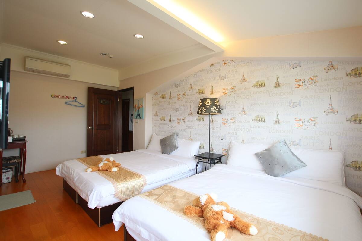 A hotel room with two beds, a nightstand, a lamp, and a wall with cityscape wallpaper.