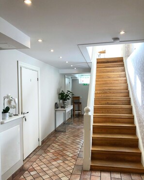 Un couloir avec un sol carrelé, un escalier en bois, une porte, une petite table avec une plante et une chaise.