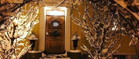 Un sendero cubierto de nieve que conduce a una puerta de madera con una luz encima, flanqueado por árboles nevados.