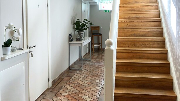 Un pasillo con escaleras de madera, paredes blancas y suelo de baldosas.