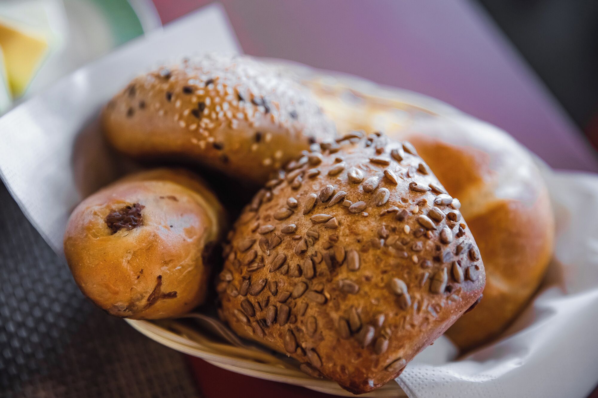 A basket of various baked goods, including bread rolls with seeds and a bun with a visible filling.