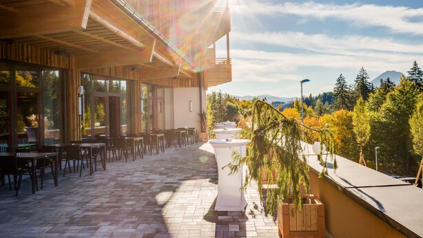 Un patio al aire libre con mesas y sillas, techo de madera y vistas a árboles y montañas al fondo.