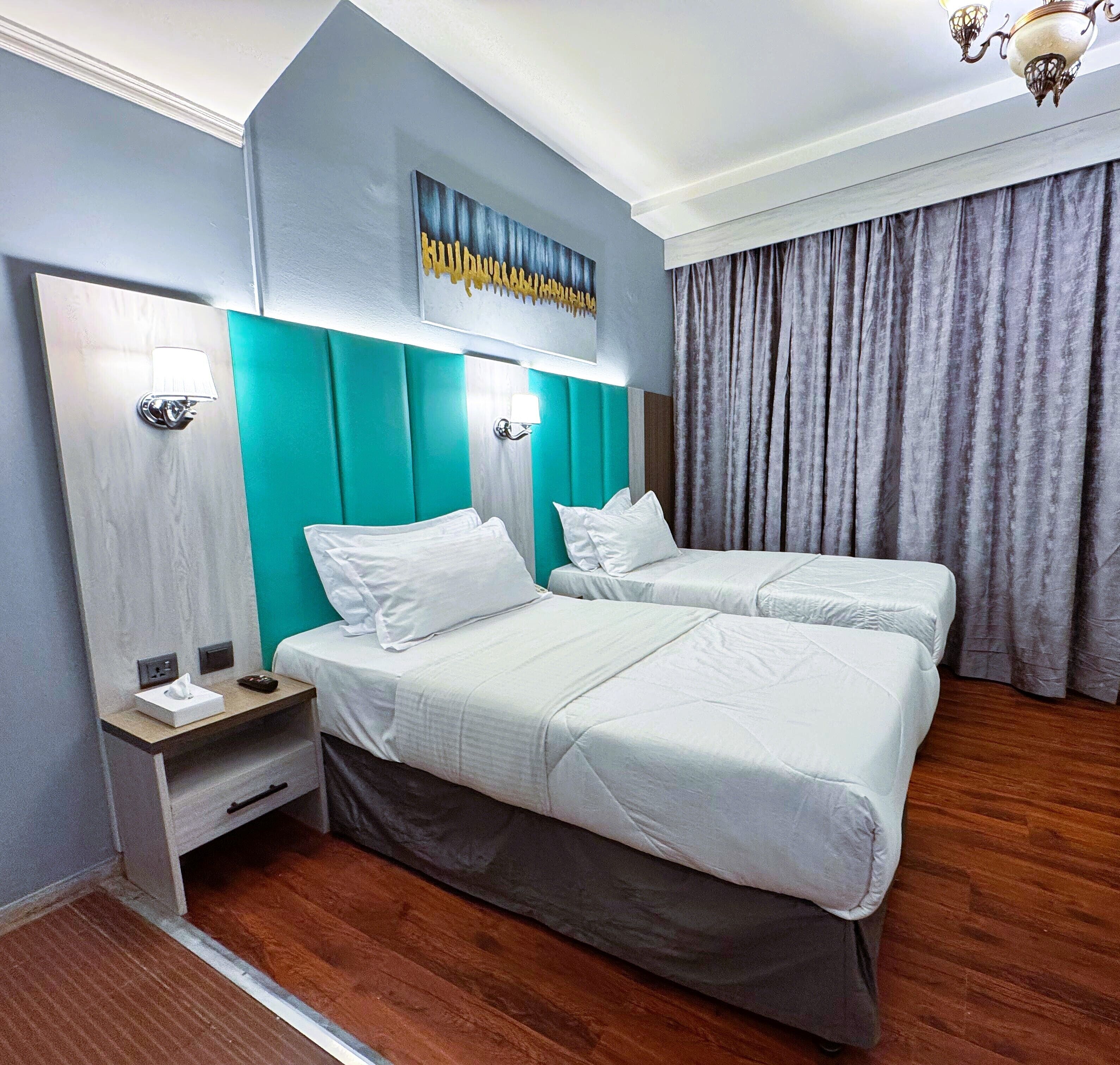 A hotel room with two beds, a teal accent wall, a chandelier, and a large window with curtains.