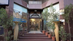 Entrada de hotel con escalones que conducen a una puerta de vidrio, rodeada de plantas en macetas y un cartel.