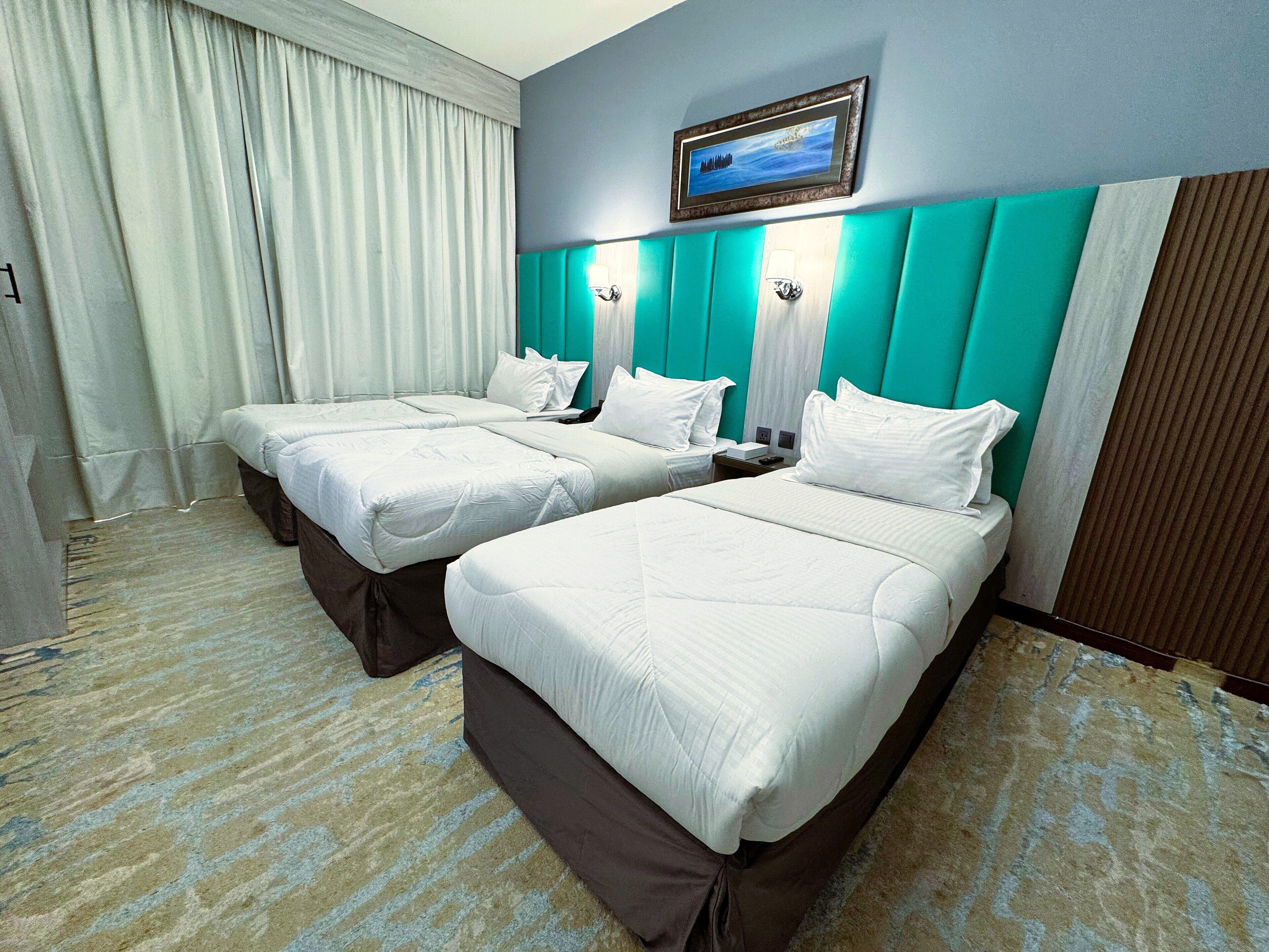 A hotel room with two beds, a large window with curtains, a painting on the wall, and a wooden headboard.