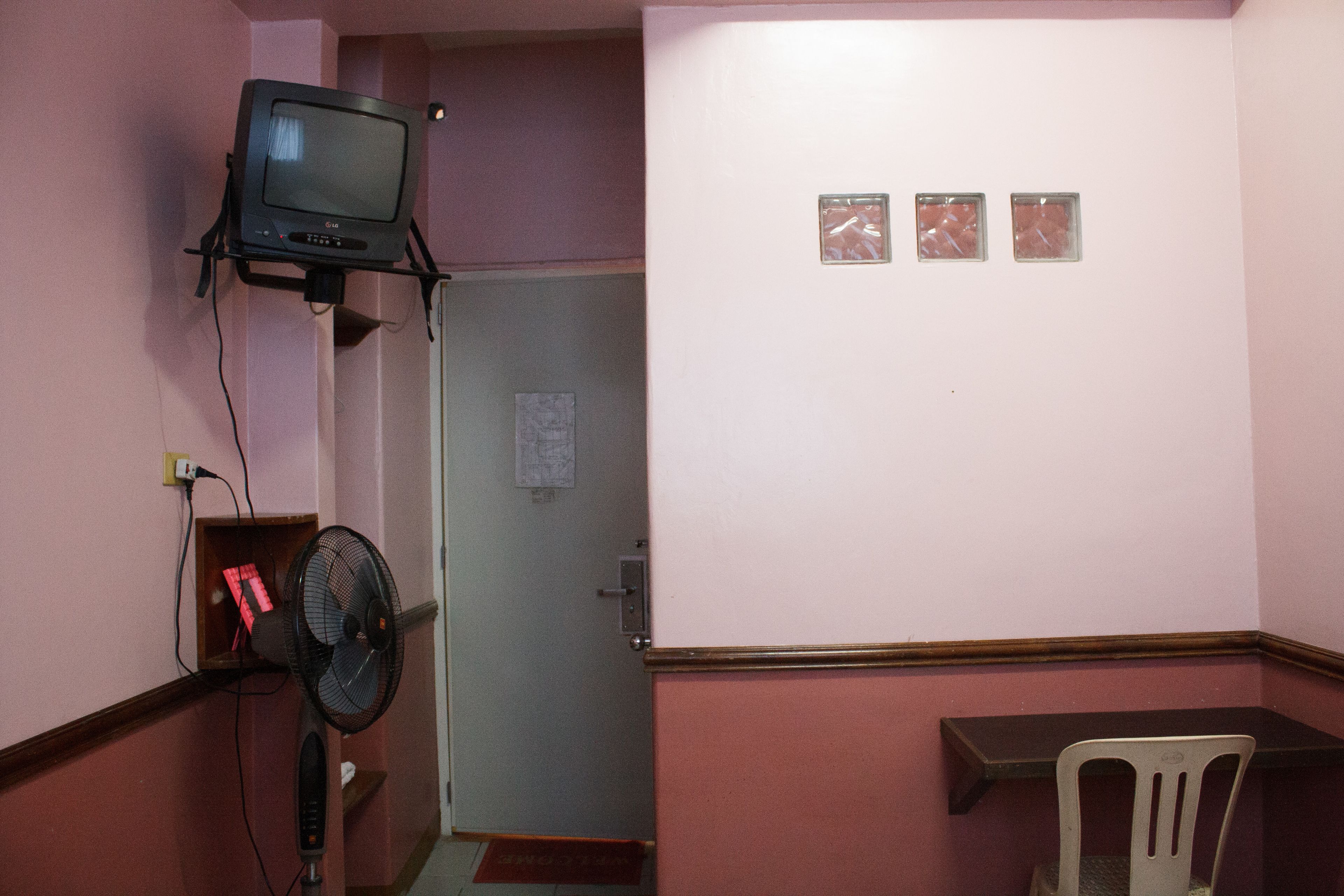 Una stanza con televisore a parete, una ventola, una porta e un piccolo tavolo con una sedia.