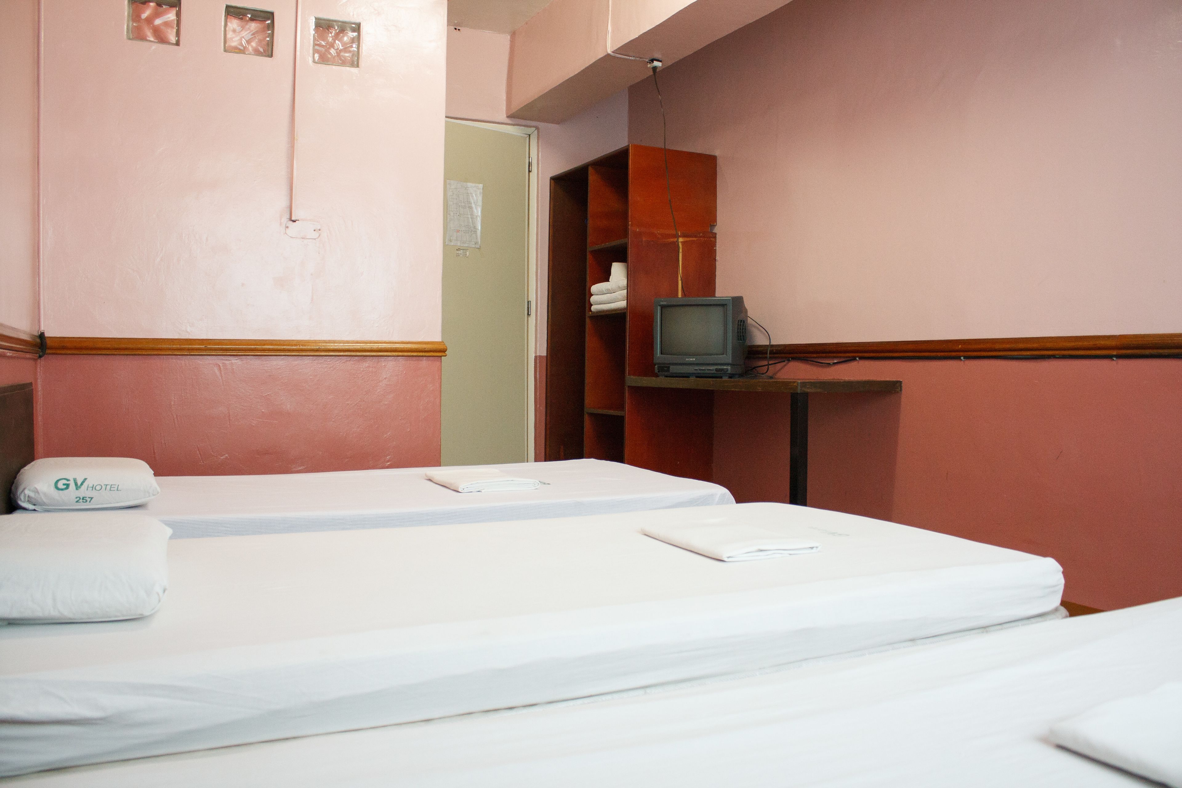 A hotel room with two beds, a television on a wooden shelf, and a door leading to another room.