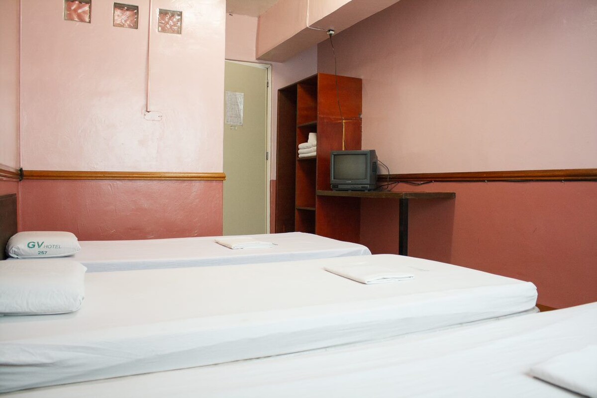 Quarto de hotel com duas camas, uma televisão sobre uma estante de madeira e uma porta que dá para outro quarto.