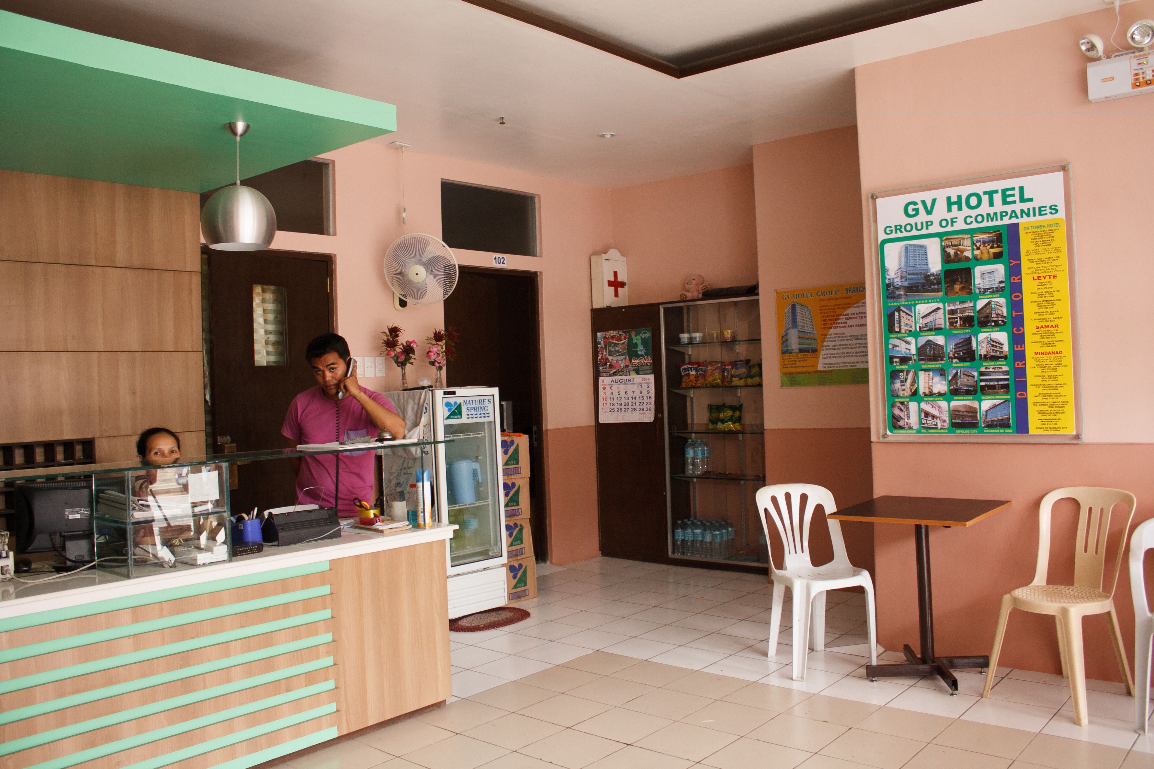 Zona de recepción de un hotel con mostrador, sillas, ventilador y un cartel en la pared.