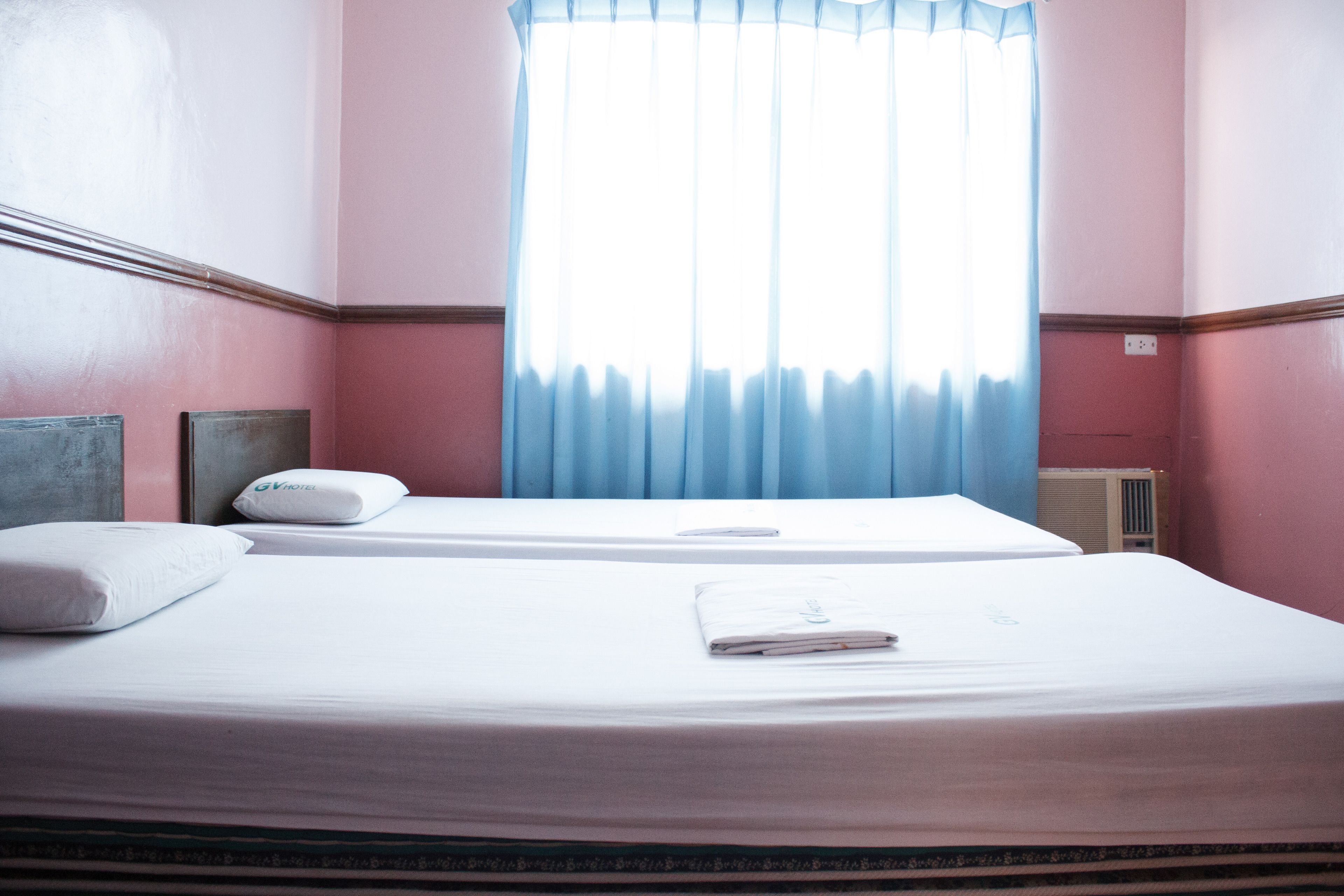 A hotel room with two beds, a window with blue curtains, and a radiator on the wall.
