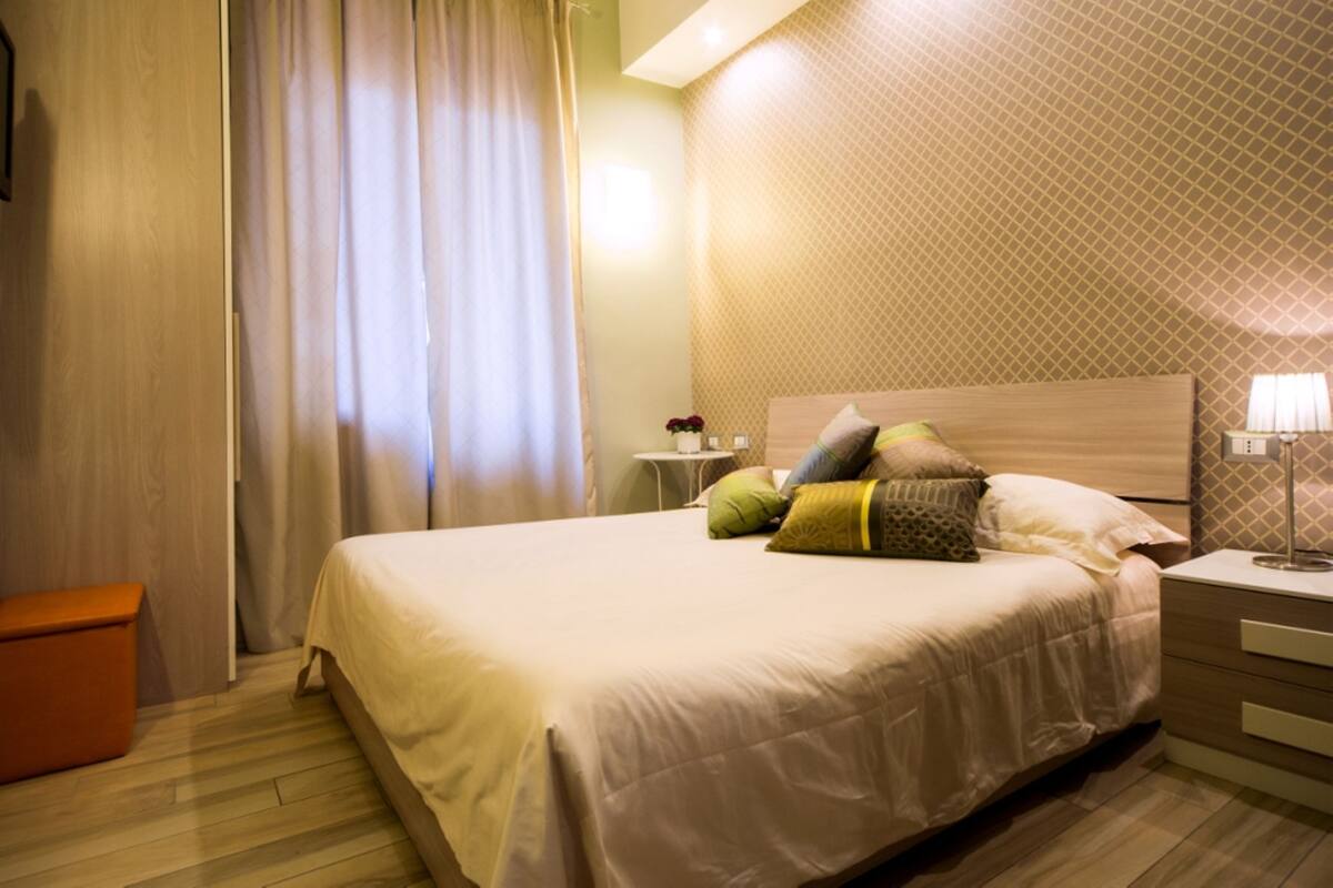 A hotel room with a large bed, a nightstand, a lamp, and a wooden floor.