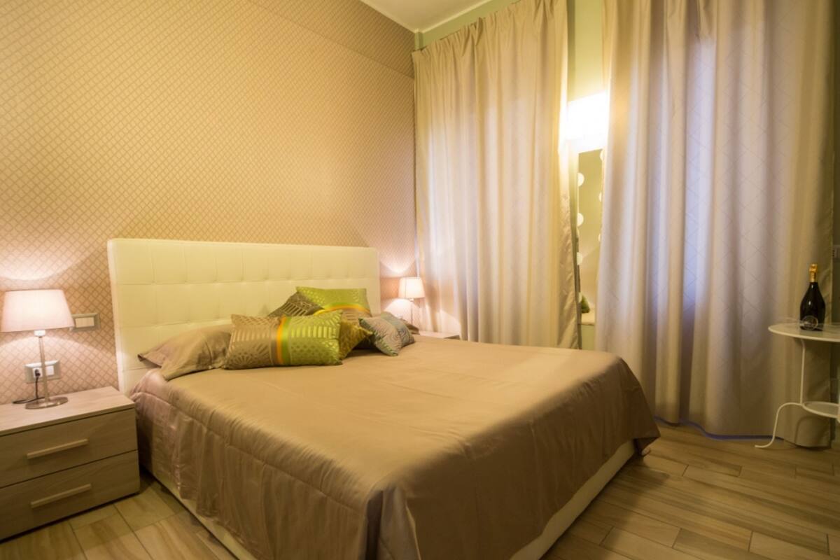 A hotel room with a large bed, a nightstand, a lamp, a bottle of champagne, and a mirror.