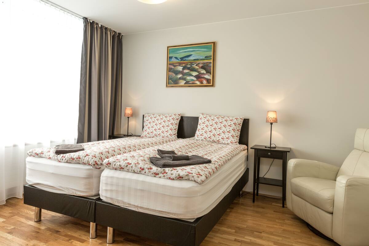 A double bed with floral bedding, a bedside table with a lamp, a chair, and a painting on the wall.