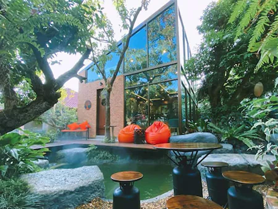 Un área de estar al aire libre con un estanque, rodeada de abundante vegetación y un edificio moderno con grandes ventanales.