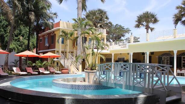 Zona de piscina con sillas de descanso y sombrillas, rodeada de palmeras y un edificio de varios pisos con balcones.